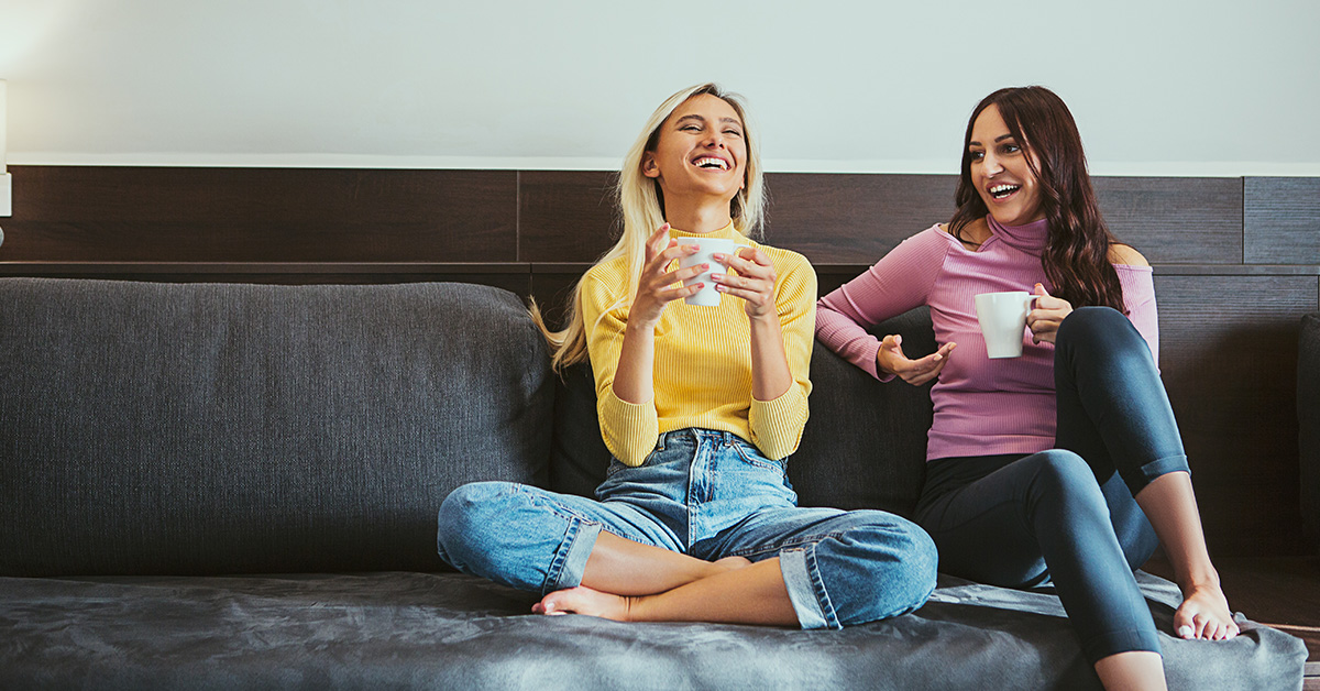 two women laughing together on couch with coffee